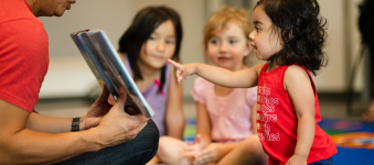 A close up of a small toddler in a reading group pointing at a book that is being read by a teacher. 