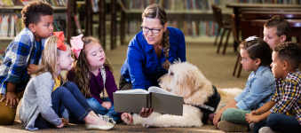 A female librarian with a fluffy service dog seated and on the floor reading to a group of small children in the library.