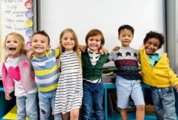 Six small kids standing with their arms around each other, smiling and laughing in the classroom. 