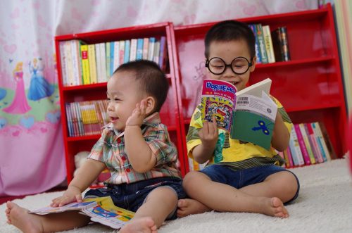 Two very young boys on the floor in a classroom reading picture books, laughing and smiling