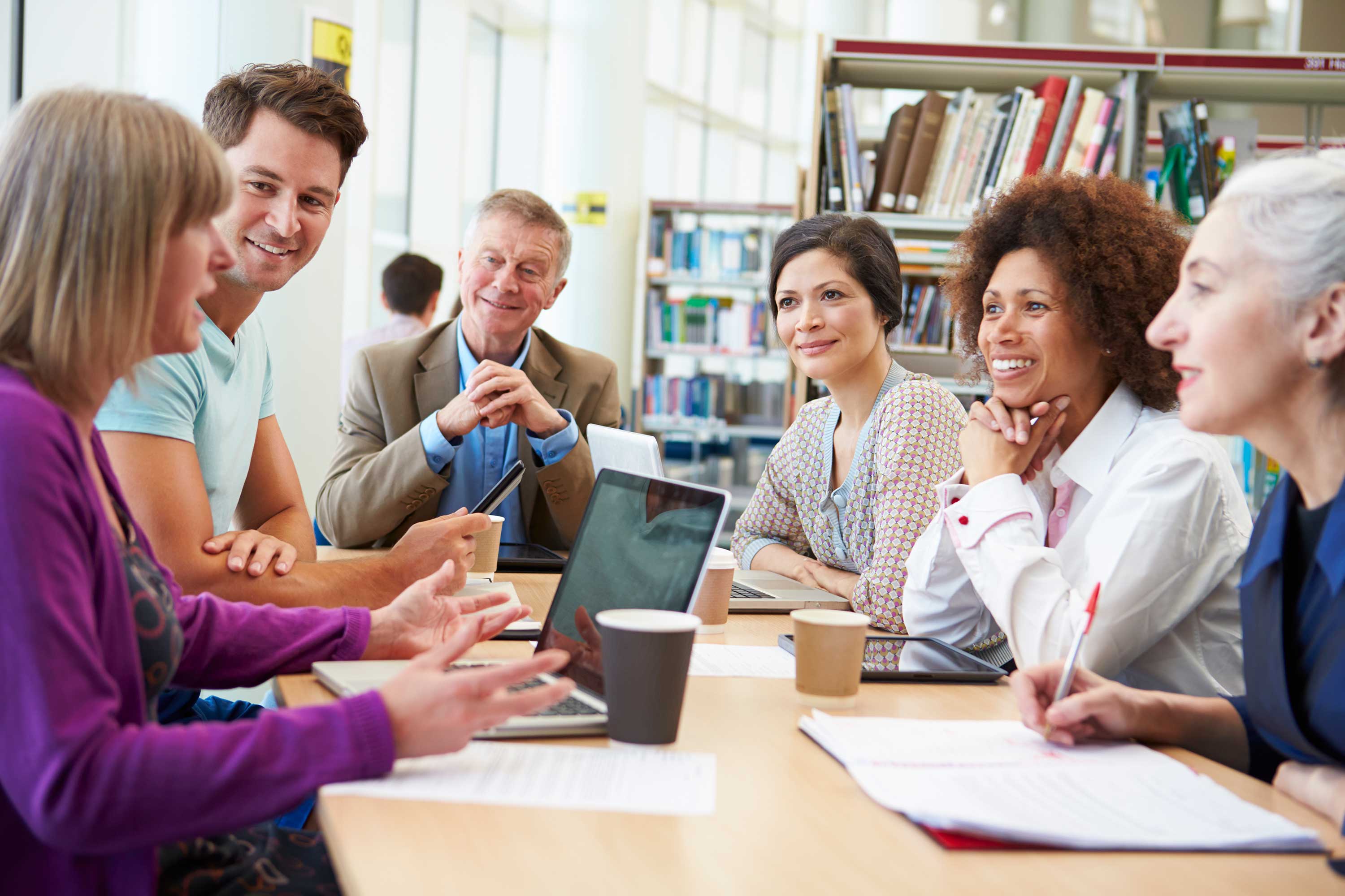 Diverse group of educators sitting around a table talking and talking notes in laptop and notebook