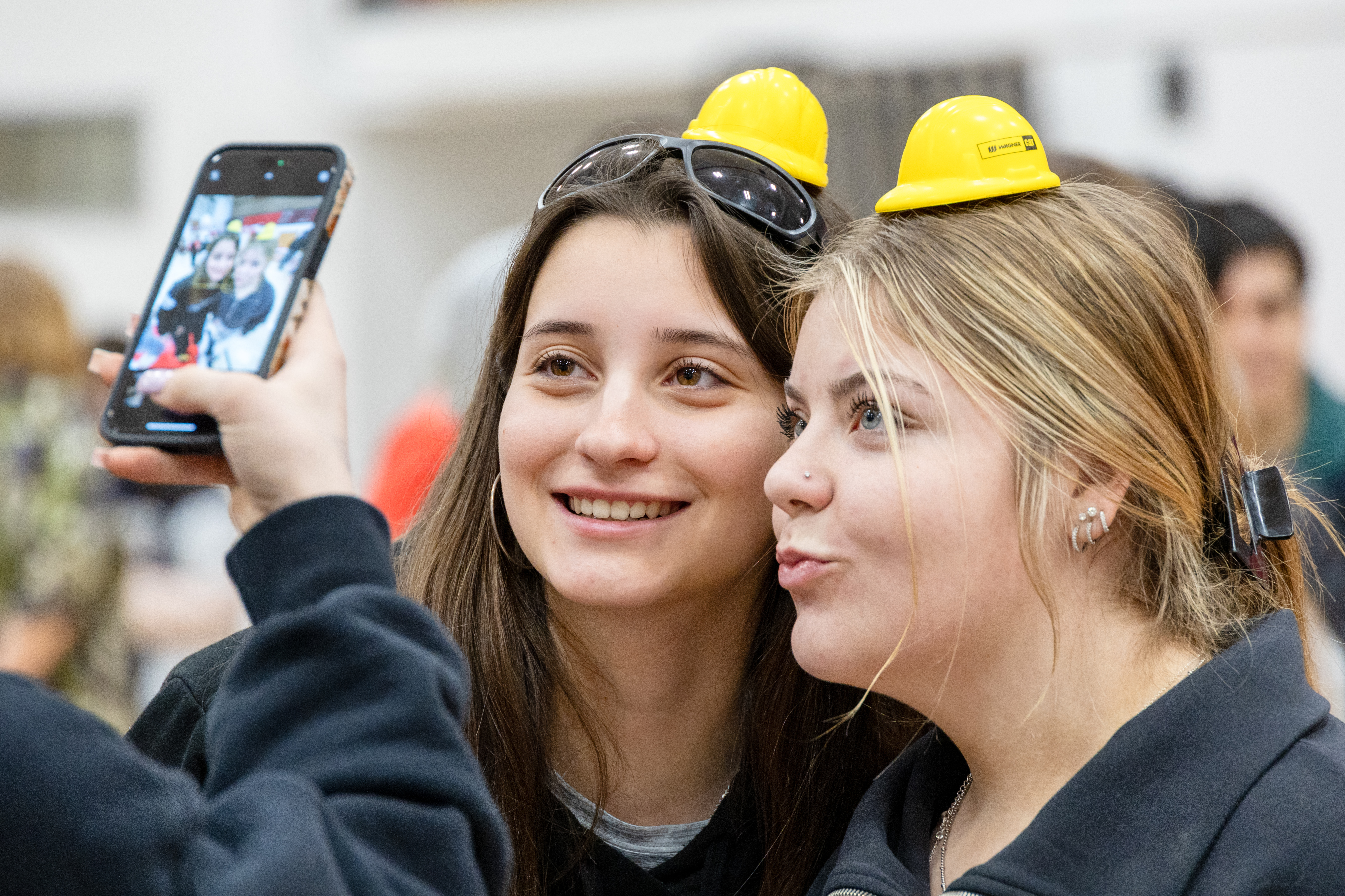 Two high school girls pose for a photo wearing tiny yellow hard hats at a career fair. 