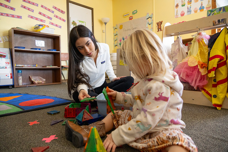A high school senior sits on the floor of a daycare with a toddler and hands her a green triangular magnetic "stick and stack" toy.