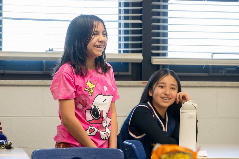 A girl wearing a pink Snoopy t-shirt stands and smiles with another smiling girl wearing a navy jersey seated next to her.