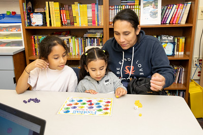 Parent with two children work on an activity at a table in an elementary school, shelves of books behind them