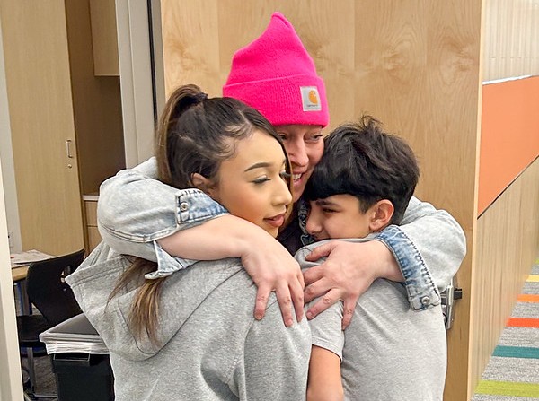 A teacher wearing a bright pink beanie hugs a tween girl and boy in front of a classroom door.