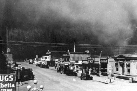 A dust storm engulfs the town of Burlington, Colorado (Kit Carson County). Cars are parked on unpaved Main Street in front of the town's commercial storefronts. A grain elevator is at the end of the street.