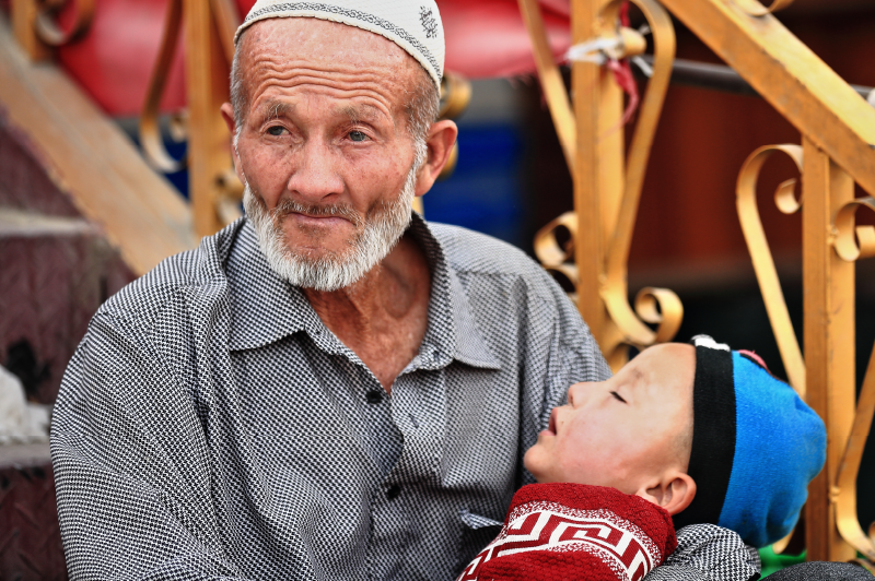 Hotan, China-October 3, 2017: Blue-eyed grandfather of the Uyghur Turkic people holds his napping grandson in his arms while they both sit on the steps of a store at the city's bazaar. Xinjiang Region