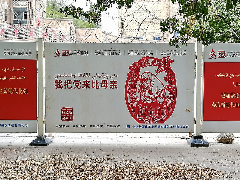 This is a roadside propaganda sign of the Communist Party of China located within the vicinity of the Turpan Museum in Turpan, Xinjiang Uygur Autonomous Region, China. The sign is written in both Uyghur and Chinese. The main message in the middle says 'I liken the party to my mother.'