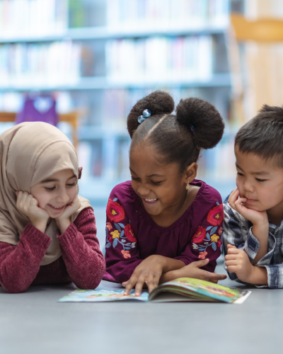 Three young children in a library setting gathered around a picture book smiling and pointing at the book