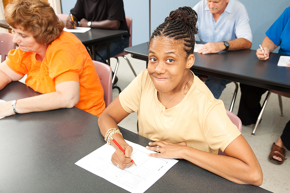 Student smiling  at a desk while writing on paper, adults at desks in background